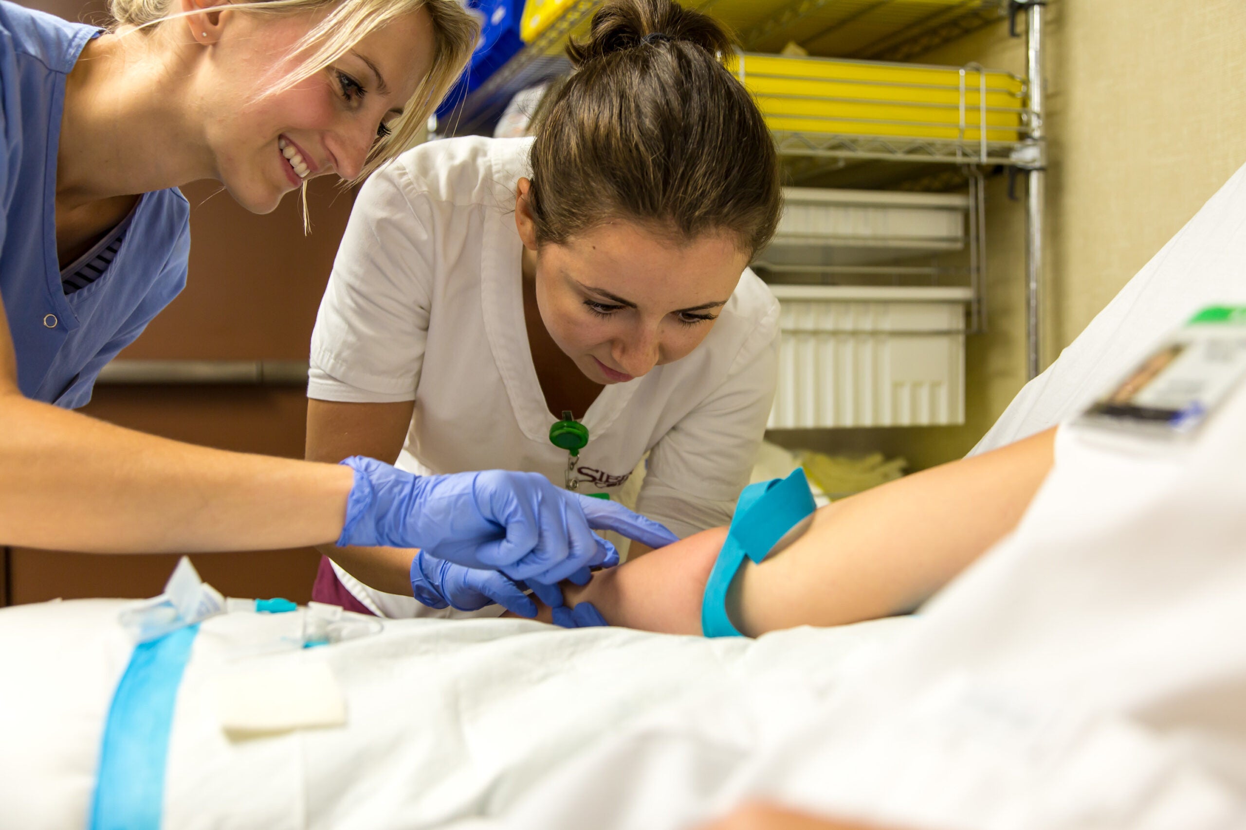 Female instructor shows female phlebotomy student how to do a blood draw on a patient's right arm