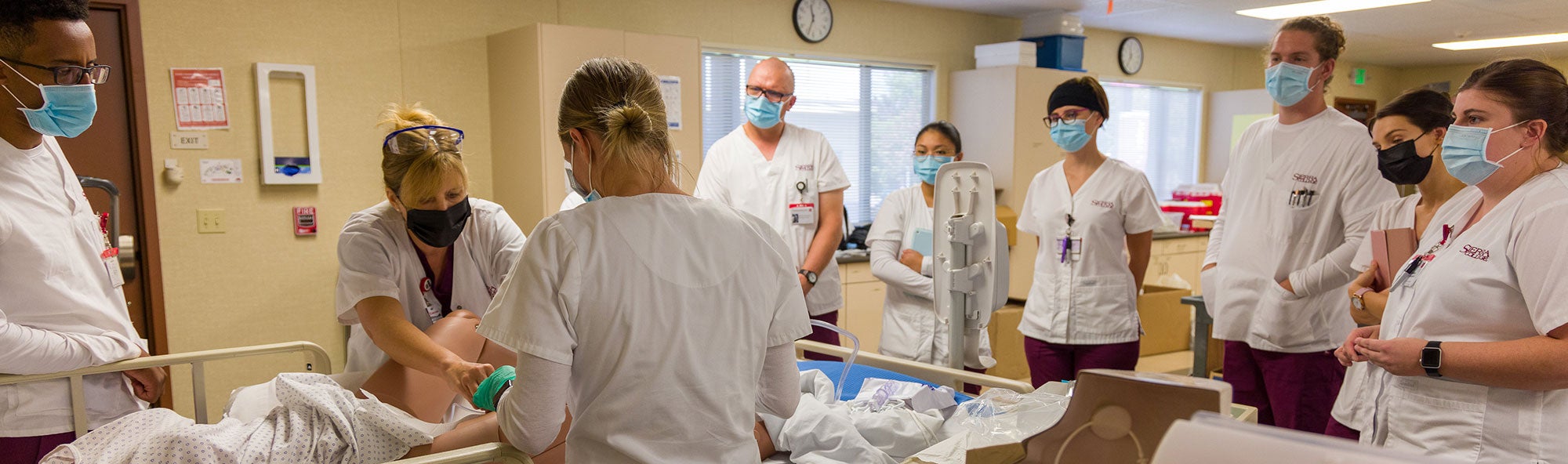 Male and female nursing students encircling a simulated patient while listening and learning from their instructor