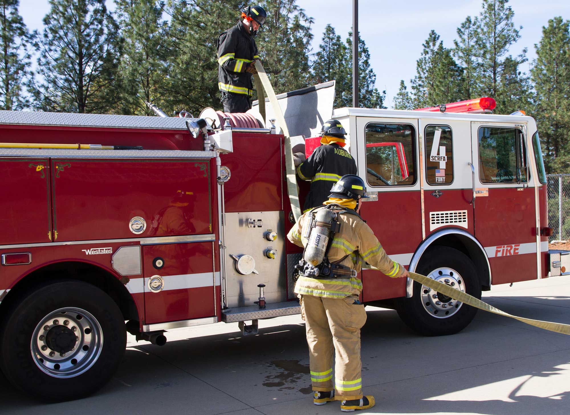 Three fire tech students wearing firefighting jacket and pants rolling up hose on red fire truck after practicing drills