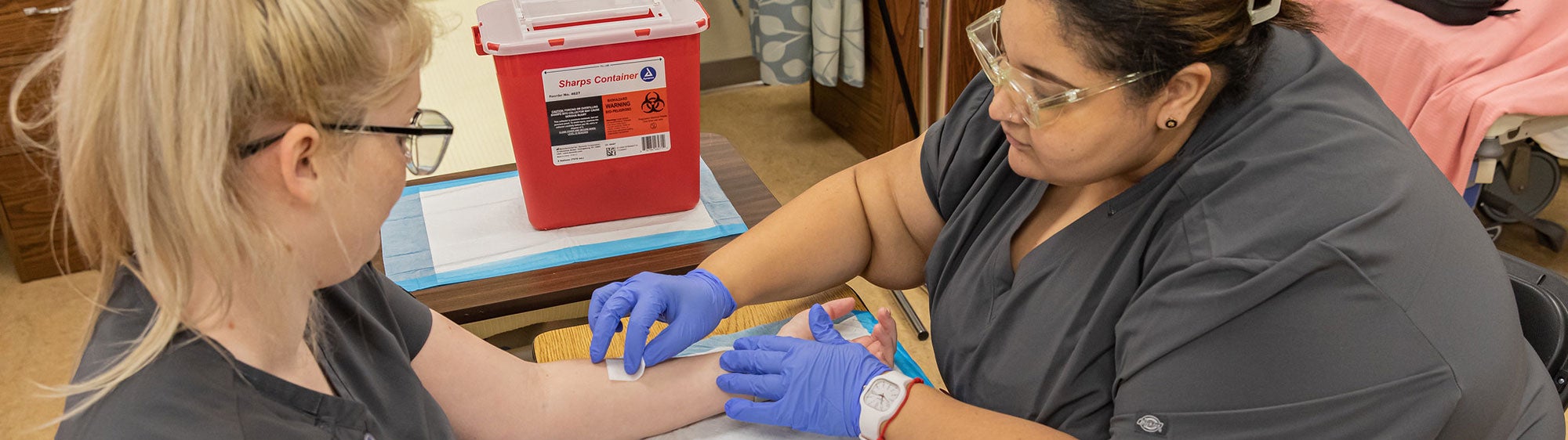 Medical Assistant students in gray scrubs practice cleaning skin before drawing blood.