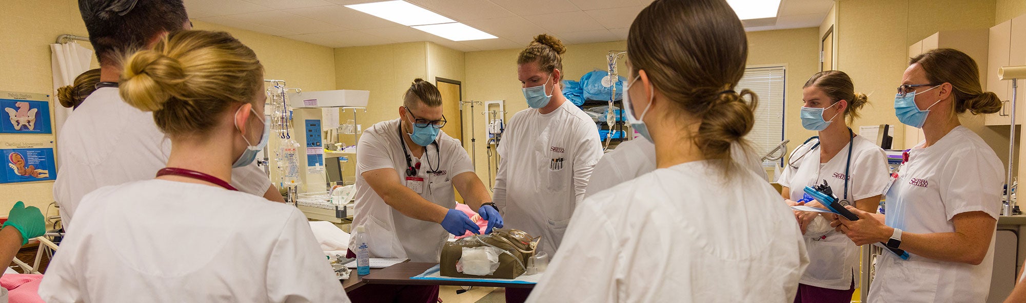 Seven students wearing scrubs while watching peers practice on simulated patient