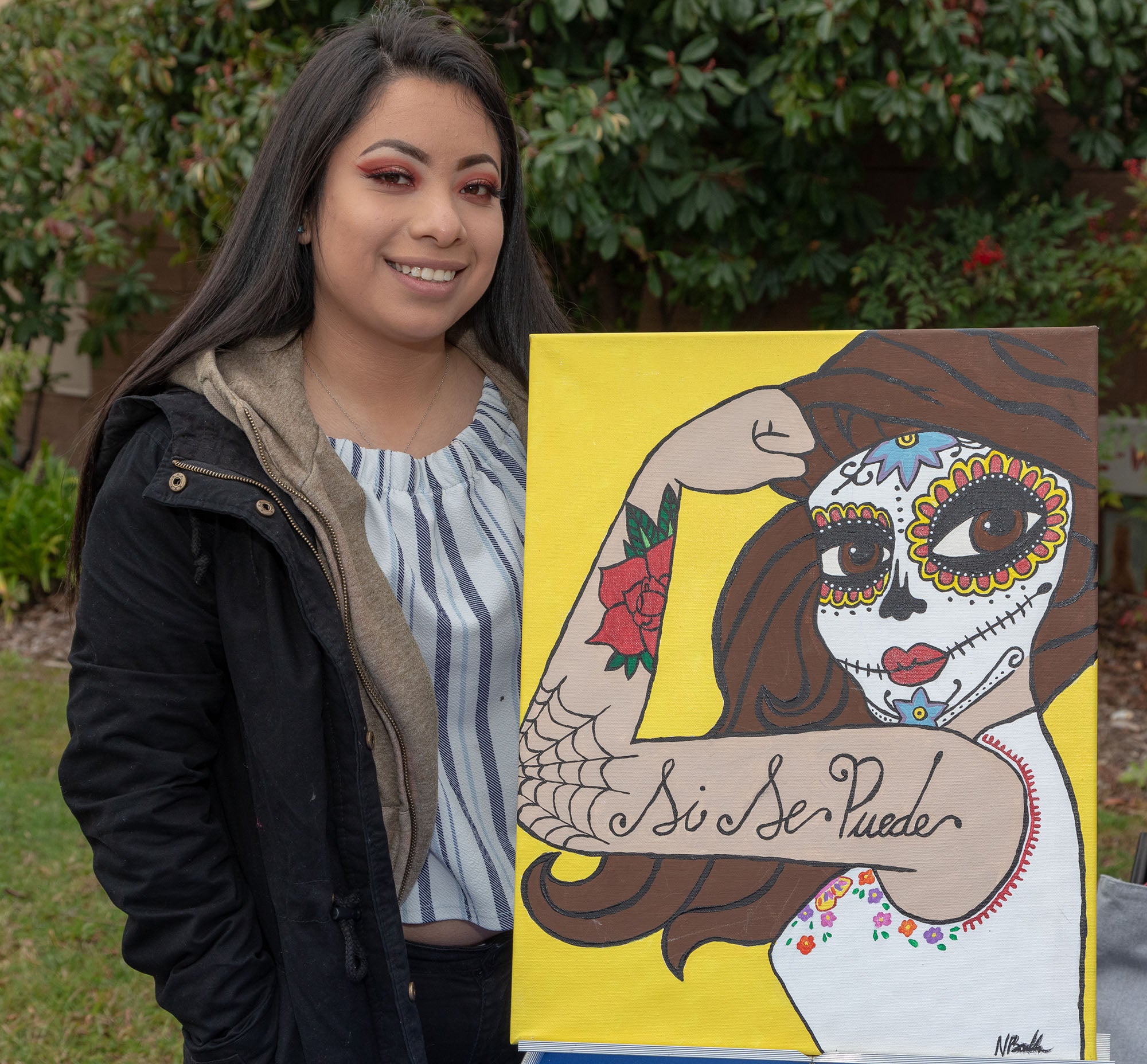 Latina student with her painting of an empowered female with calavera makeup and the words si se puede