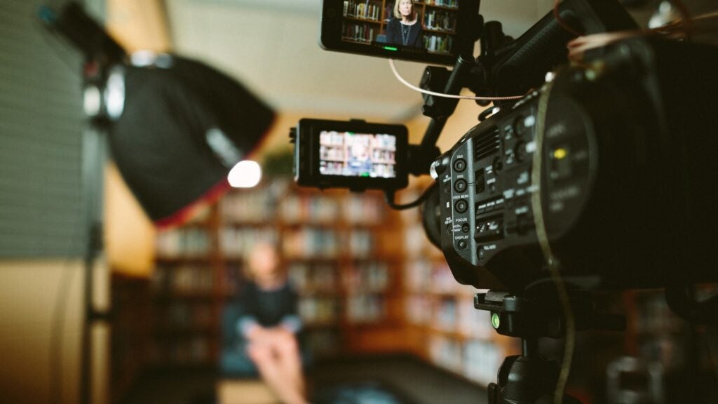 Video camera pointing at interviewee sitting in chair with bookcase in background - Photo by Sam McGhee on Unsplash