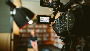 Video camera pointing at interviewee sitting in chair with bookcase in background