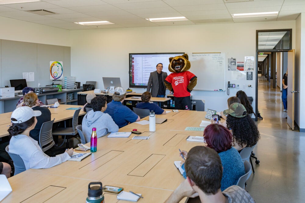 New Sierra College mascot, wearing a red shirt and black shorts, stands in front of classroom full of students, alongside instructor.
