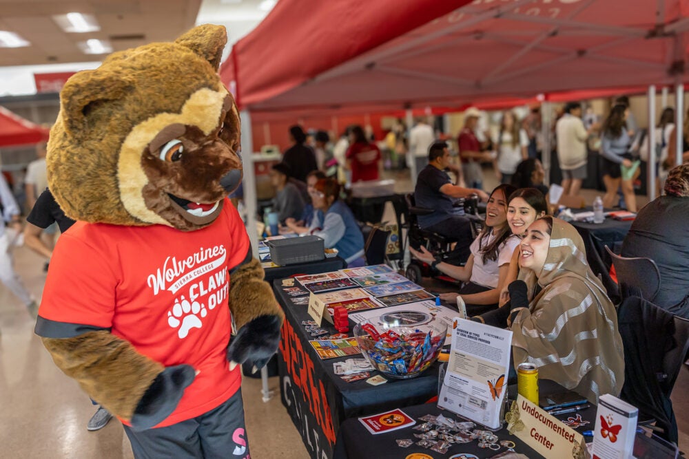 New Sierra College mascot, wearing a red shirt and black shorts, looking at informational tables with fliers and swag on the table, employees behind the table are smiling at the mascot