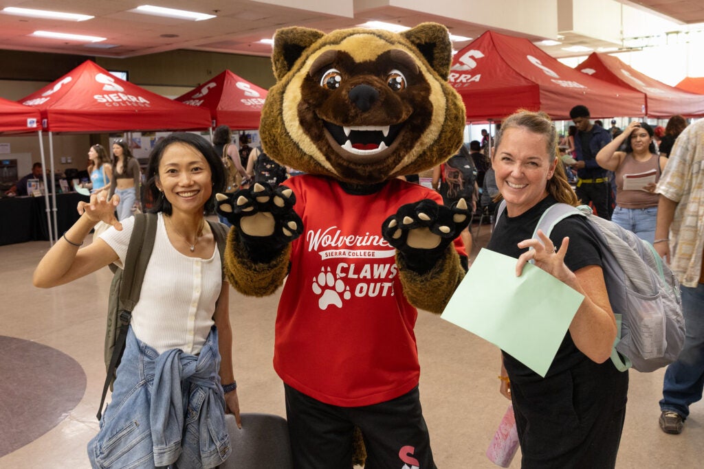 Sierra College Wolverine Mascot, wearing a red shirt and black shorts, poses with two students. All have their hands up like claws.