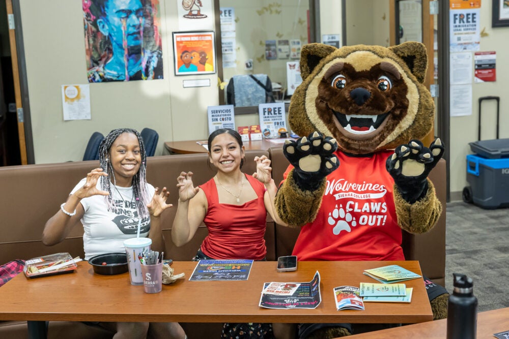 New Sierra College mascot, wearing a red shirt and black shorts, poses with students on campus, holding their hands up in the shape of claws.