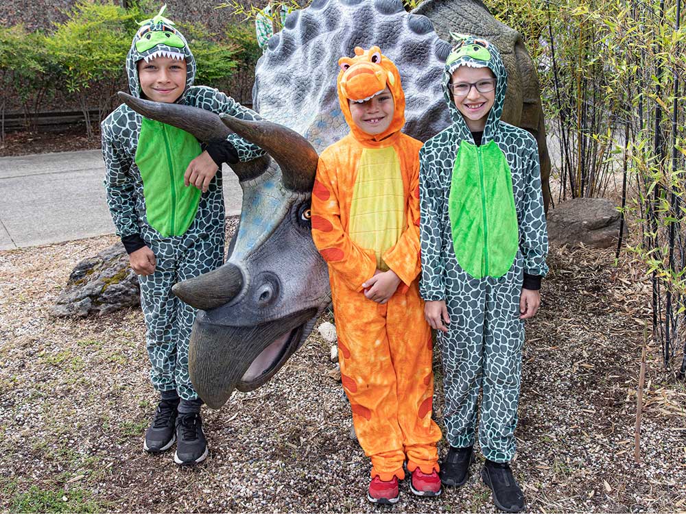 Three kids in dinosaur costumes standing next to dinosaur