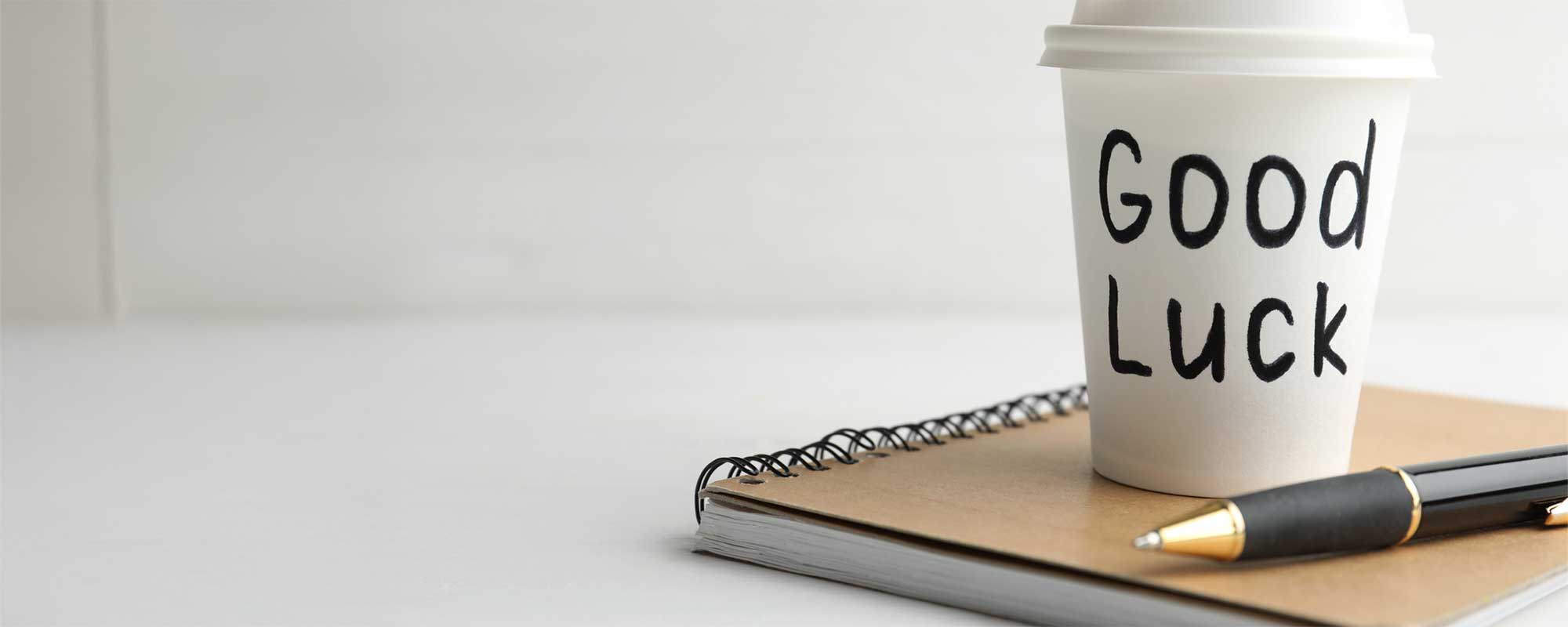 Coffee cup says Good Luck sitting on notepad with pen