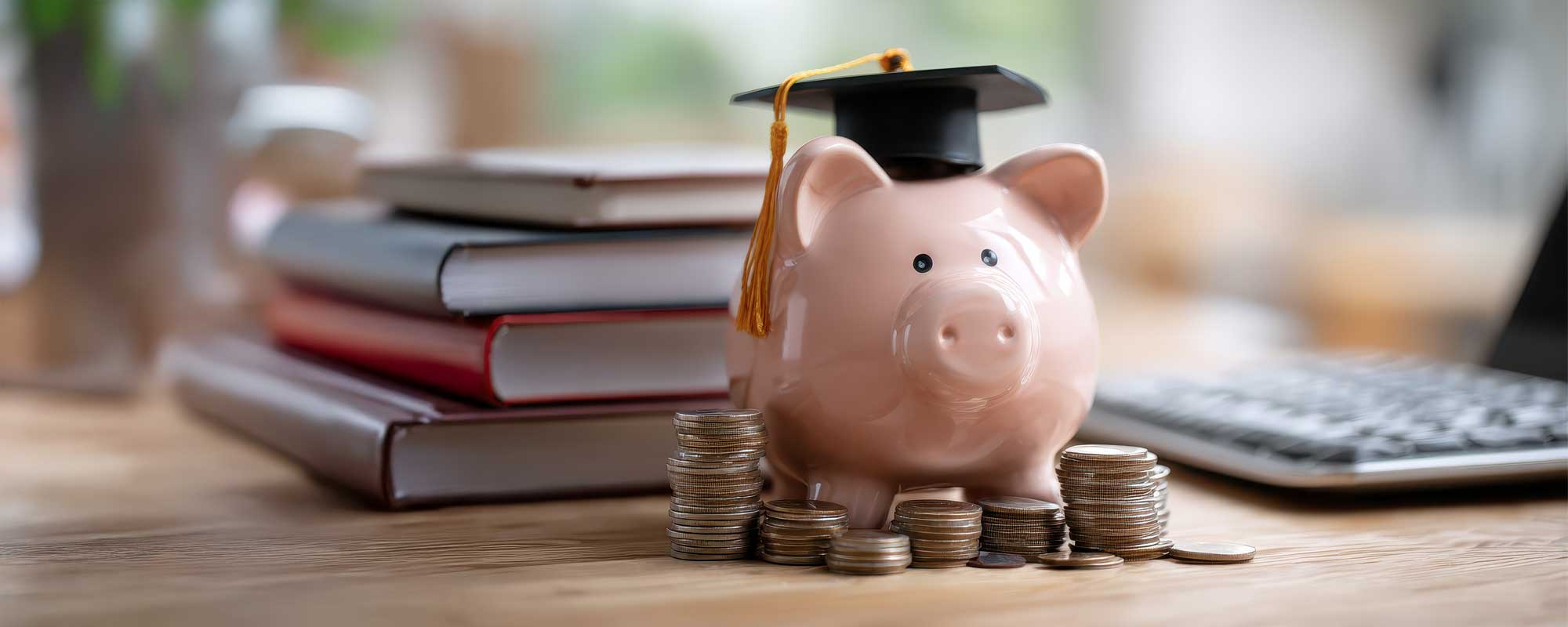 Piggy bank wearing graduation cap next to stack of coins, books and laptop on desk