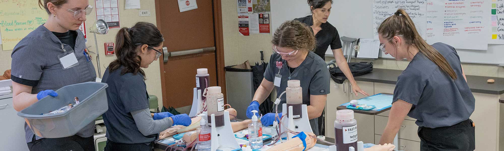 Medical Assistant students in gray scrubs practice cleaning skin before drawing blood.